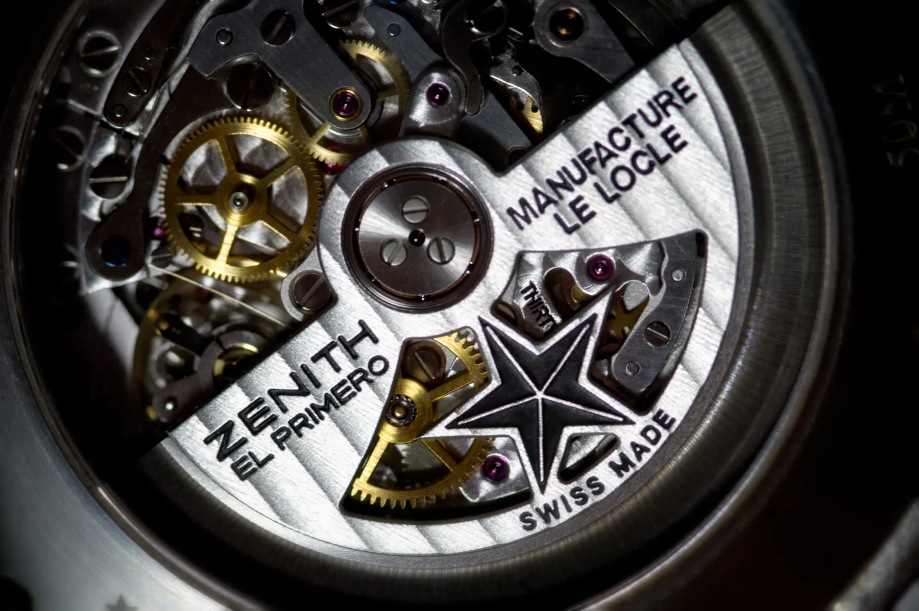 Close-up photograph through a caseback of a Zenith El Primero automatic chronograph movement, showing the brushed steel rotor engraved with the Zenith star logo and the words ZENITH EL PRIMERO, MANUFACTURE LE LOCLE and SWISS MADE, with gold-coloured gear wheels and pink jewels visible through the rotor's cut-out