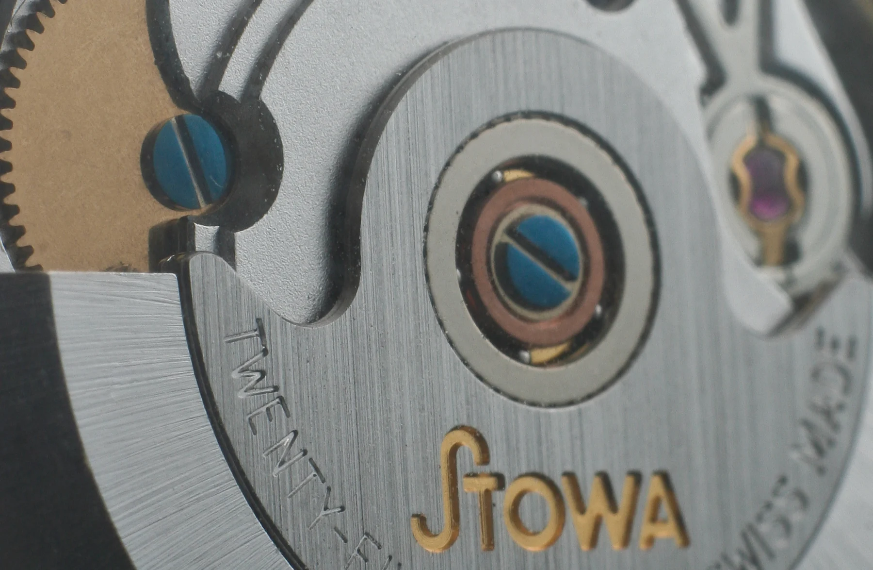 Macro photograph through a watch caseback of an ETA 2824 automatic movement in a Stowa Antea, showing the brushed steel rotor engraved 'TWENTY-FIVE 25 JEWELS SWISS MADE', its central ball-bearing pivot, blued screws, a gold-coloured gear wheel at the left edge and a pink jewel set in a gold chaton at the right