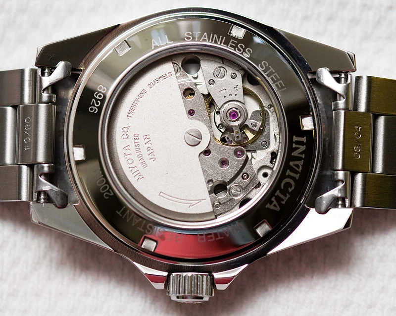 Photograph through the caseback of an Invicta 8926 dive watch showing a Miyota 8215 automatic movement: a plain grey plate engraved 'MIYOTA CO. UNADJUSTED JAPAN' and 'TWENTY-ONE JEWELS', with the balance wheel and pink jewels visible on the right and a curved arrow marking the rotor's winding direction