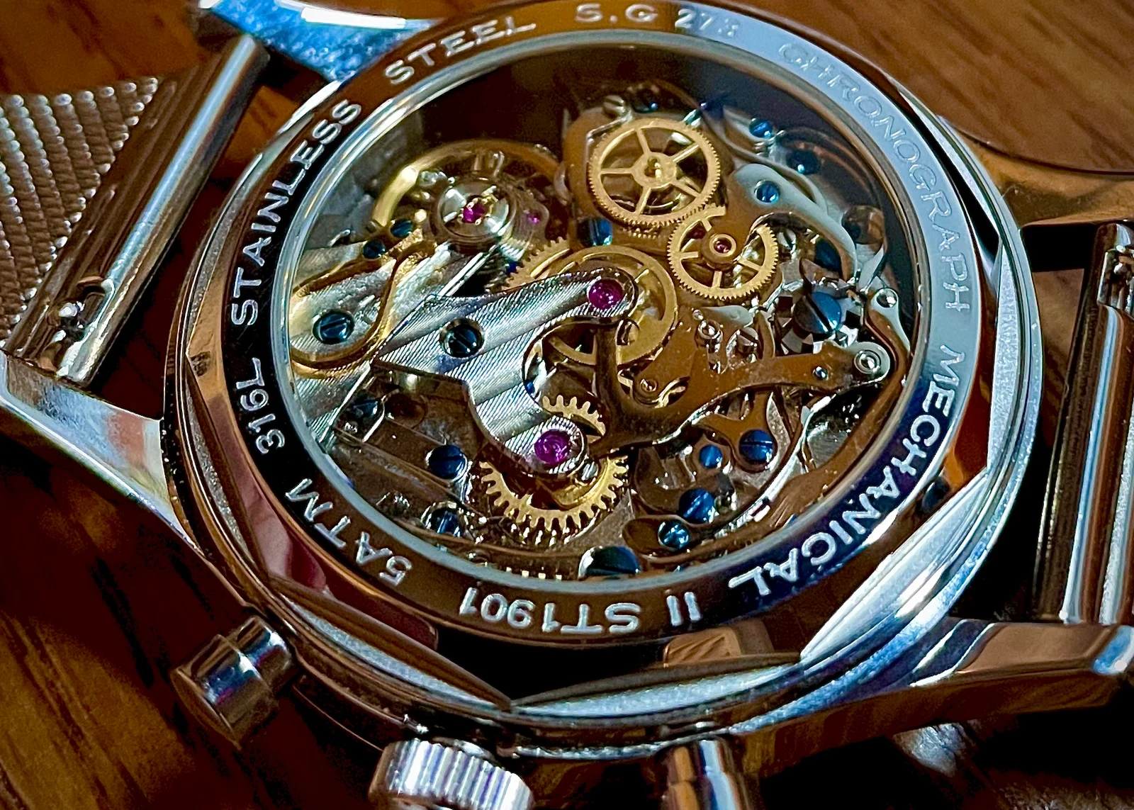 Photograph of a stainless steel chronograph watch lying face-down on a wooden surface, its exhibition caseback engraved '316L STAINLESS STEEL' and 'MECHANICAL CHRONOGRAPH ST1901', revealing a gold-plated hand-wound movement with a prominent column wheel, curved steel chronograph levers, blued screws and several pink jewels