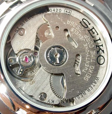 Photograph through a watch caseback of a Seiko 7S26 automatic movement, showing a plain brushed steel plate engraved 'SEIKO TIME CORP.' and 'TWENTY-ONE JEWELS 7S26A', with the balance wheel and a pink jewel visible at the left and the winding train at the top — an undecorated, purely functional movement