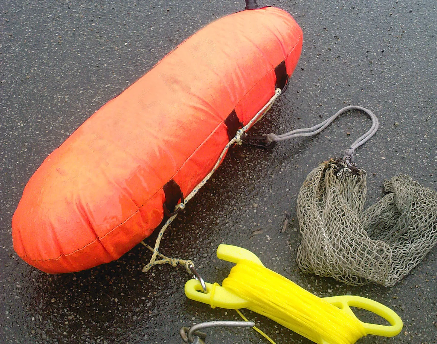 Photograph of a deflated orange inflatable torpedo tow buoy lying on wet asphalt. Four black webbing loops are stitched along its flank with a light line threaded through them, and beside it lie a mesh catch bag and a yellow plastic line winder with line wound around it.